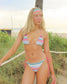 Woman in a colorful striped bikini standing on a beach with grass and a palm tree in the background.