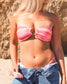 Woman wearing a pink striped bikini top with blue jeans against a stone wall.