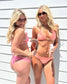 Two women wearing pink and peach bikini sets pose together against a white slatted wall, holding drinks in a sunny outdoor setting.