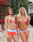Two women in matching red and blue bikinis standing on a beach with plants and flowers in the background.