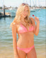 A woman wearing a pink two-piece bikini stands on a beach with water and boats in the background, holding a small red lollipop.