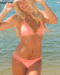 A woman wearing a light pink lace-trimmed bikini and a beige straw hat stands on a sunny beach with water and boats in the background.
